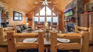 Een knus cabin interieur met houten wanden, een open haard, een televisie en een eethoek met houten stoelen en een tafel voor vier personen.