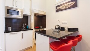 Cocina compacta con gabinetes blancos, encimera negra y microondas. Hay una pequeña isla con tapa de cristal y taburetes rojos. En la pared, encima de la isla, hay un cuadro de un pez.