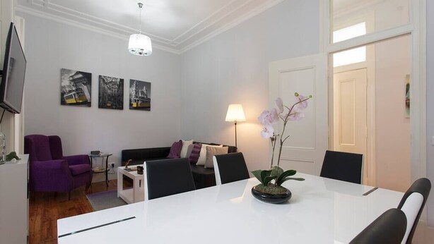 Un salón moderno con una mesa de comedor blanca, sillones morados, un sofá negro y un televisor a la izquierda.