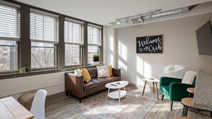 Una sala de estar moderna con un sofá de cuero marrón, un sillón verde, una mesa de centro blanca y una alfombra estampada. Hay ventanas grandes con persianas, un letrero en la pared y una rejilla de ventilación en el techo.