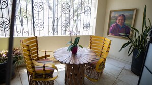 A room with two yellow chairs and a round table with a white floral pattern. There is a potted plant on the table, and a painting on the wall.