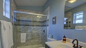 A bathroom with a glass shower enclosure, a toilet, a sink with a mirror, and a brown rug on the floor. - Oceanfront Luxury Apartment Overlooking Kachemak Bay and Homer Spit! (Homer)