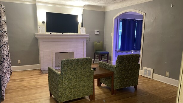 Een woonkamer met een open haard, twee gestreepe fauteuils, een houten salontafel en een flatscreen televisie die boven de open haard is bevestigd.