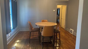 Een eetgedeelte met een ronde tafel en vier stoelen, een raam met gordijnen, en een deur naar een andere kamer.