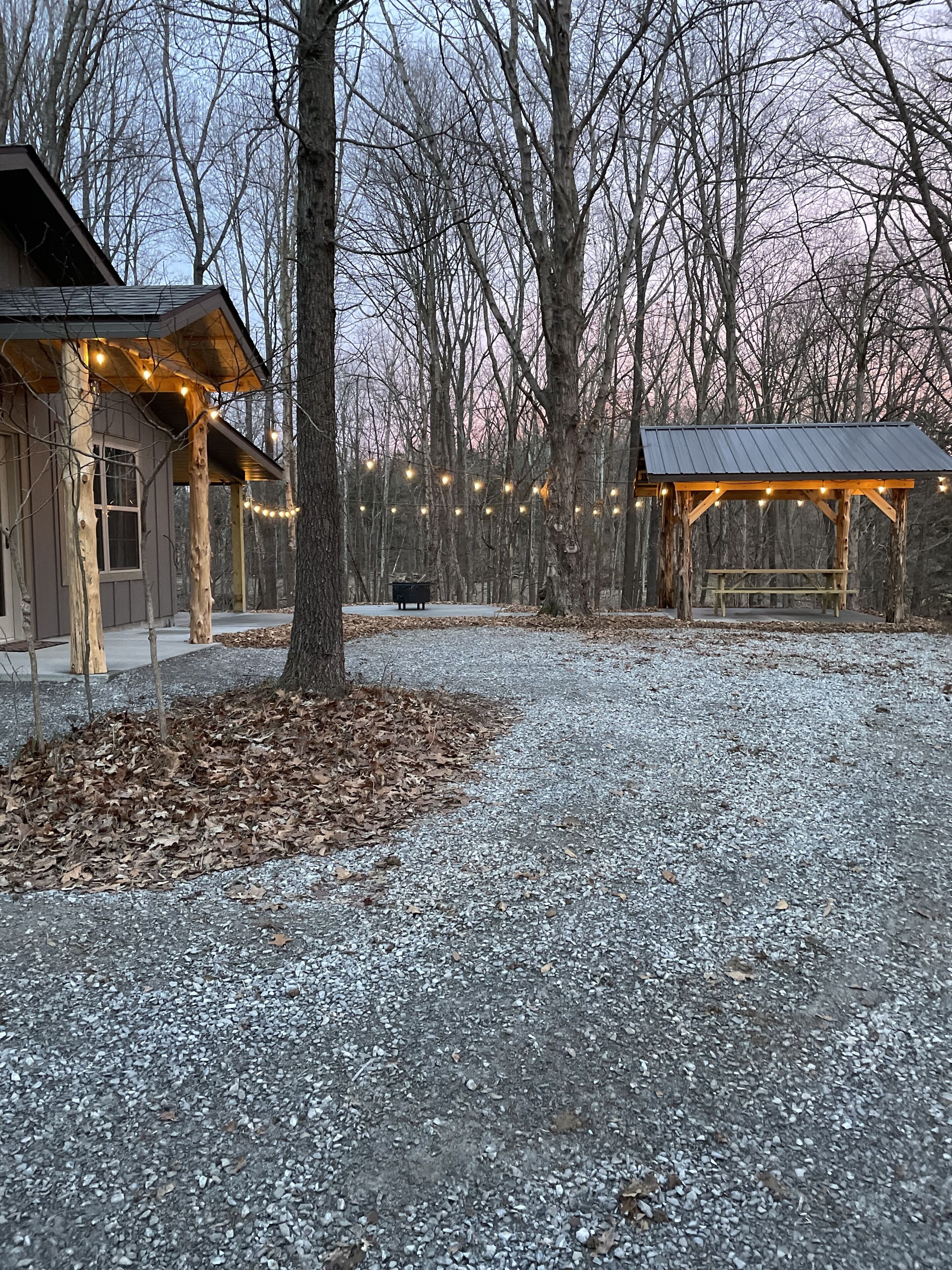 A rustic cabin with a covered porch and a picnic area, surrounded by trees and gravel paths.
