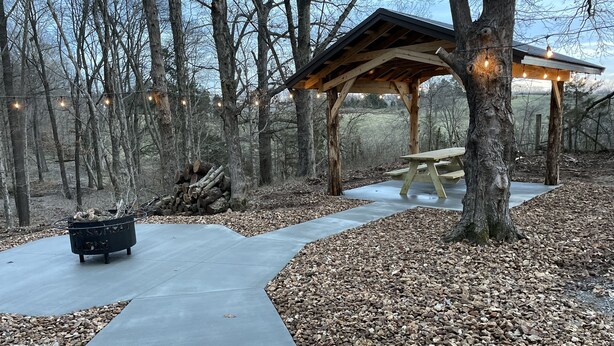 A wooden gazebo with string lights, a picnic table, and a fire pit in a forested area.
