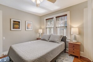 Une chambre à coucher comprenant un lit, deux tables de chevet avec des lampes, une fenêtre avec des stores et deux œuvres d’art encadrées accrochées au mur.