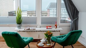 Une pièce moderne avec deux fauteuils verts, un radiateur blanc, une fenêtre donnant sur des immeubles, une plante, une bouteille, des verres et un plateau avec une théière et une tasse.