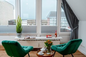 Een moderne kamer met twee groene stoelen, een witte radiator, een raam met uitzicht op gebouwen, een plant, een fles, glazen en een dienblad met een theepot en kopje.