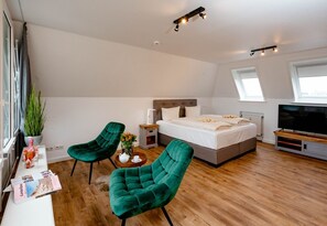 Ein modernes Schlafzimmer mit einem großen Bett, zwei grünen Sesseln, einem Fernseher und einem kleinen Tisch mit Blumen.