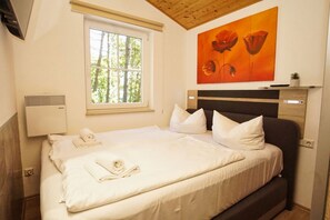 A neatly made bed with white linens, a window with a view of trees, a painting of flowers on the wall, and a wall-mounted air conditioner. - Chalet Ostsee No. 72 - chic vacation bungalow close to the beach with terrace - (Göhren)