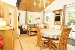 A cozy living room with a sofa, a dining table with chairs, a wooden ceiling, and large windows.
