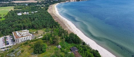 An aerial view of a coastal area with a beach, a large building with multiple floors, and a parking lot.