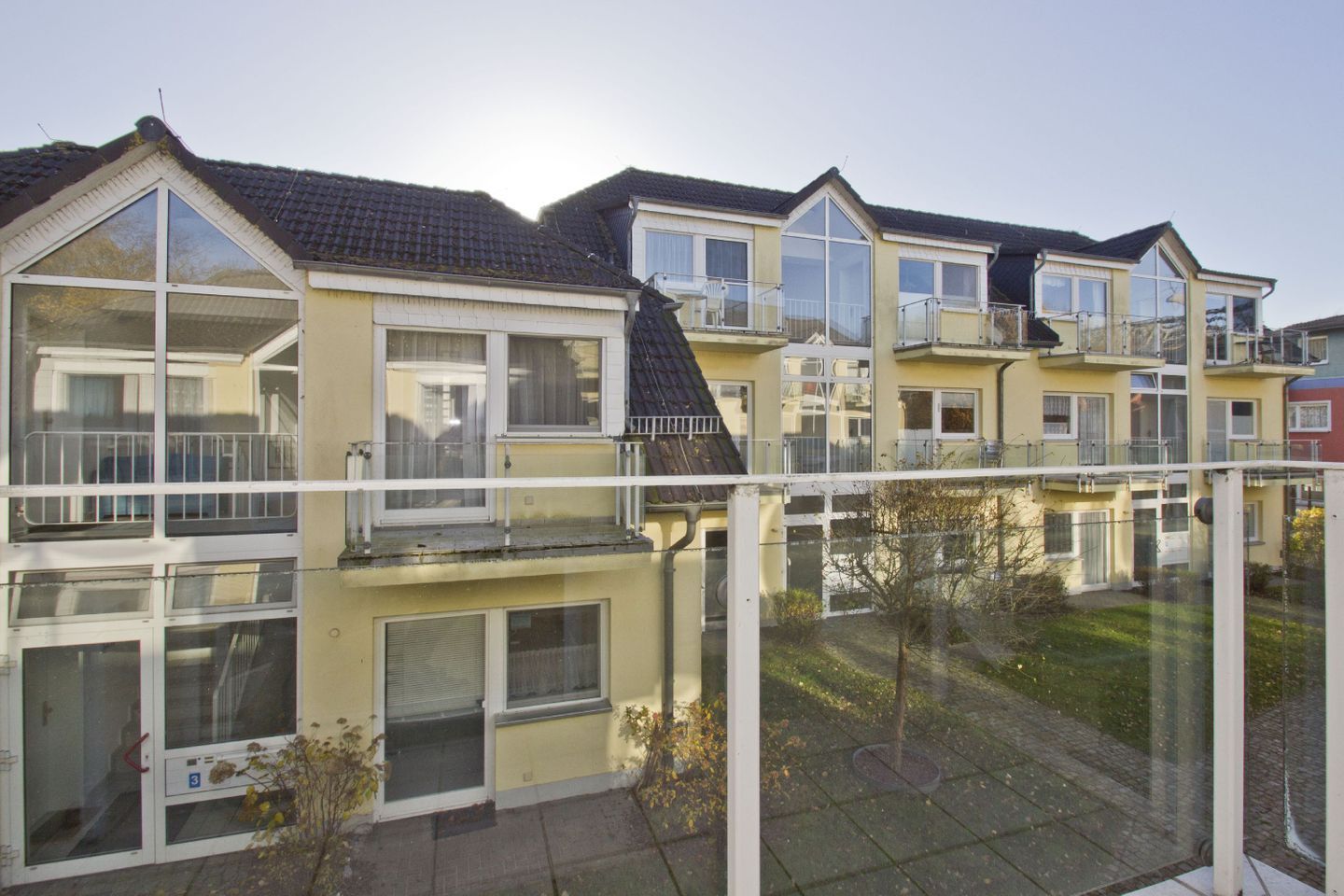 A row of yellow residential buildings with large windows and balconies, a fenced yard, and a clear sky.