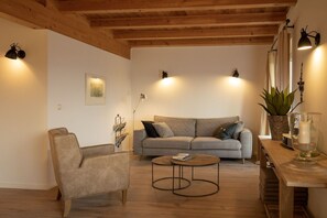 Un salon chaleureux avec un canapé, un fauteuil et une table basse, agrémenté de poutres en bois au plafond et d’un éclairage fixé au mur.