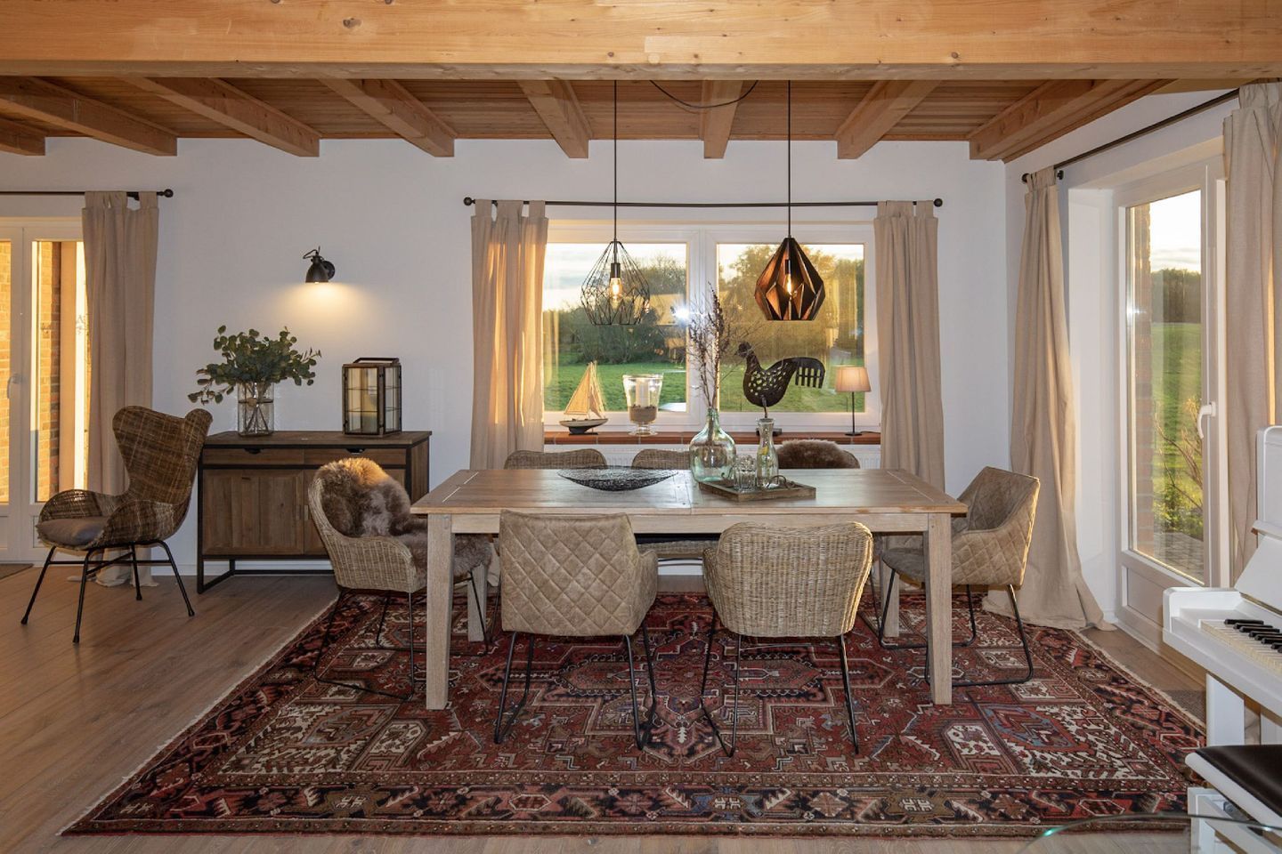 Un coin repas avec une table en bois, des chaises en osier, un tapis à motifs et une vue sur la verdure à l’extérieur.