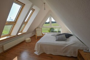 A bedroom with a sloped ceiling, skylights, a bed with pillows, a chair, and a view of a green landscape.
