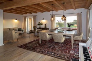 A spacious living room with a wooden ceiling, a dining area with a large table and chairs, a piano, and a view of the outdoors through a window.