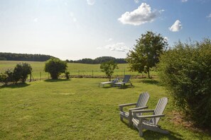 A grassy outdoor area with Adirondack chairs and a small table, surrounded by trees and a fence.