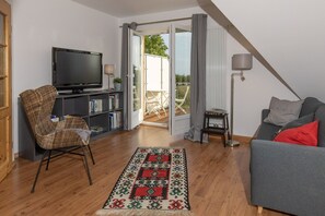 Un salon comprenant un téléviseur, une bibliothèque, un canapé gris avec des coussins rouges, un tapis à motifs et une petite table avec une lampe.