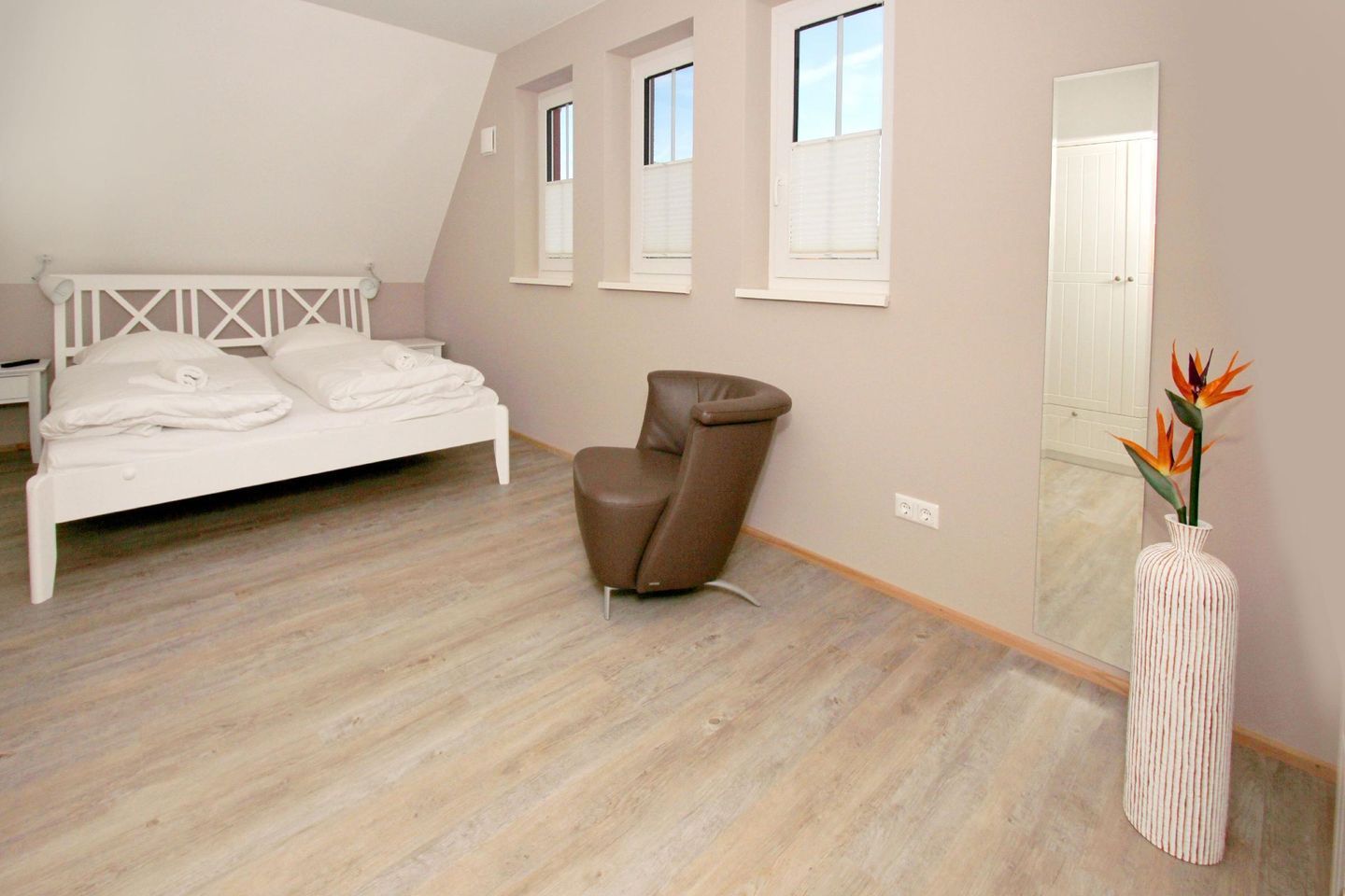 A bedroom with a bed, a brown chair, a mirror, and a vase with flowers.