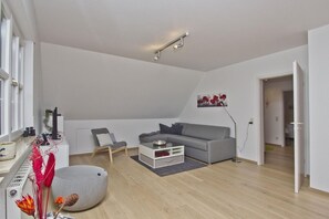 En moderne stue med en grå sofa, et lille bord og en stol. Der er en radiator, en plante og et billede på væggen.