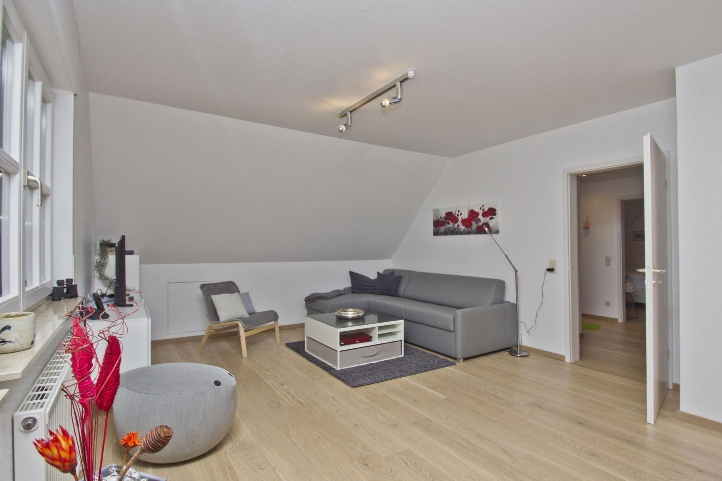 Een moderne woonkamer met een grijze bank, een klein tafeltje en een stoel. Er is een radiator, een plant en een schilderij aan de muur.