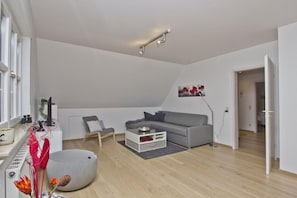 A modern living room with a grey sofa, a small table, and a chair. There is a radiator, a plant, and a painting on the wall. - Beachfront vacation apartment with a beautiful garden - Ferienhaus Gisela - Vacation Home (Putbus)
