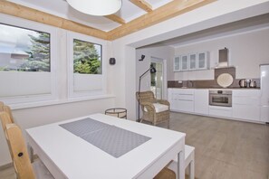 A modern kitchen with white cabinetry, a built-in oven, and a dining area with a white table and wicker chair.