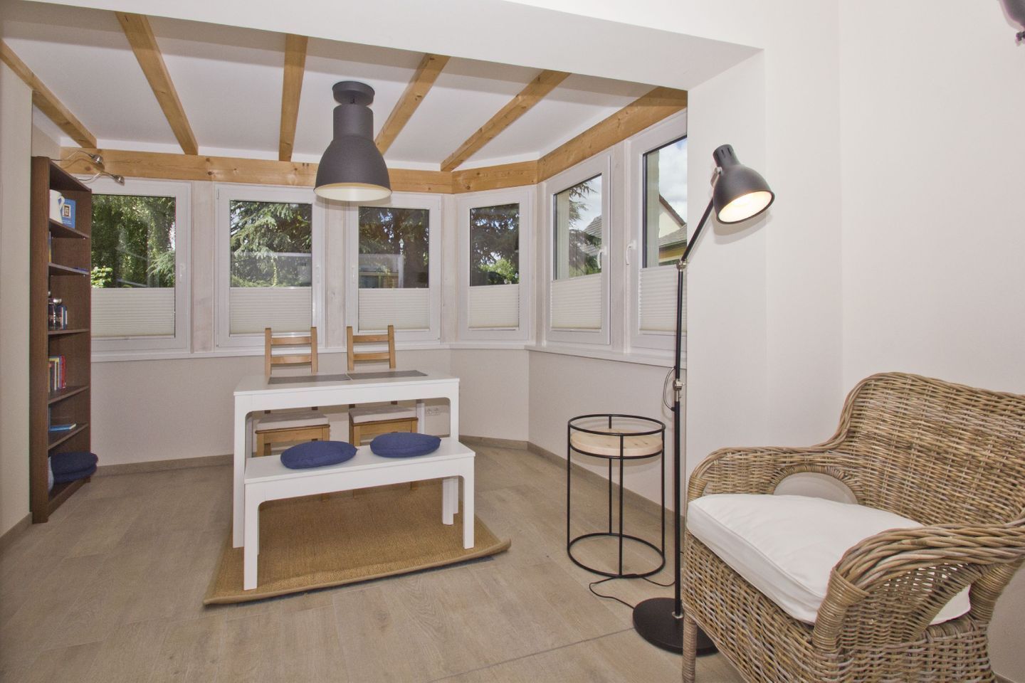 Une pièce avec une table blanche, des chaises, une bibliothèque, un canapé en osier et un lampadaire.