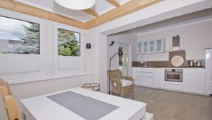 A modern kitchen with white cabinetry, a built-in oven, and a dining area with a white table and wicker chair. - Quiet location close to the sea, terrace and garden - Ferienhaus Auszeit FeWo 01 - (Middelhagen)