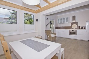 A modern kitchen with white cabinetry, a built-in oven, and a dining area with a white table and wicker chair. - Quiet location close to the sea, terrace and garden - Ferienhaus Auszeit FeWo 01 - (Middelhagen)