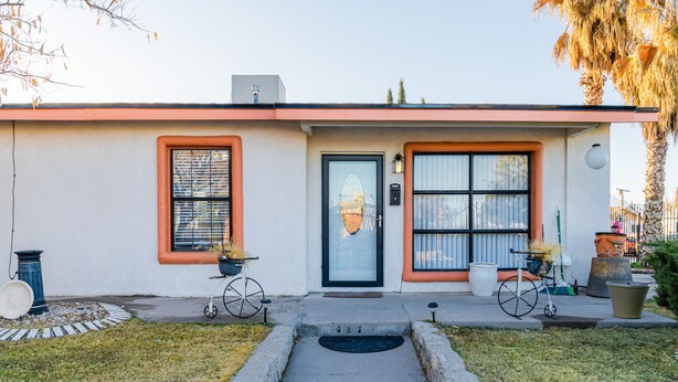 Een gelijkvloers huis met een voordeur, twee ramen met luiken, een klein portiek en een betonnen pad dat naar de deur leidt.