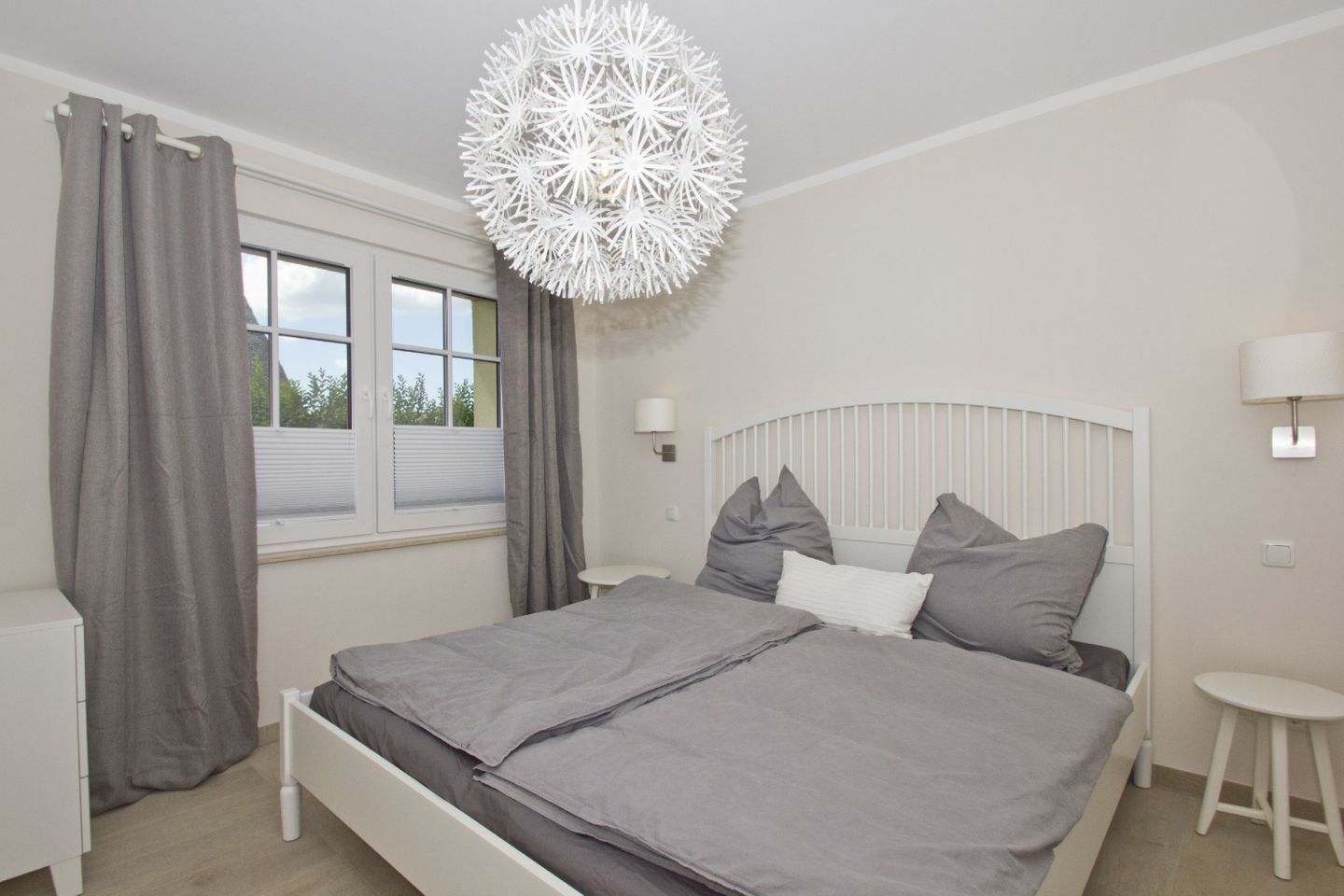 Une chambre à coucher avec un lit, des rideaux, une fenêtre, une tête de lit blanche, une table de chevet et une lampe fixée au mur.