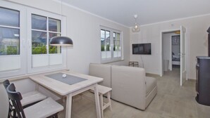 Una sala de estar moderna con un sofá, una mesa de comedor con sillas, un televisor montado en la pared y ventanas grandes con persianas.