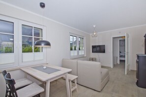 A modern living room with a sofa, a dining table with chairs, a TV mounted on the wall, and large windows with blinds.