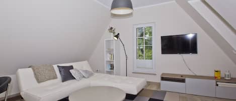 Un salon moderne avec un canapé blanc, une table ronde, une bibliothèque, un téléviseur et une grande fenêtre.