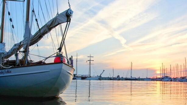 Un velero amarrado en un puerto deportivo, con aguas tranquilas y una puesta de sol al fondo.