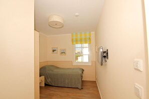 A small, narrow room with a bed, a window with a checkered curtain, a wall-mounted TV, and a light fixture.