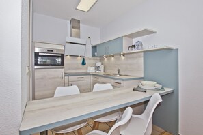 Cocina compacta con horno integrado, cafetera y lavabo. Hay dos sillas blancas y una mesita.