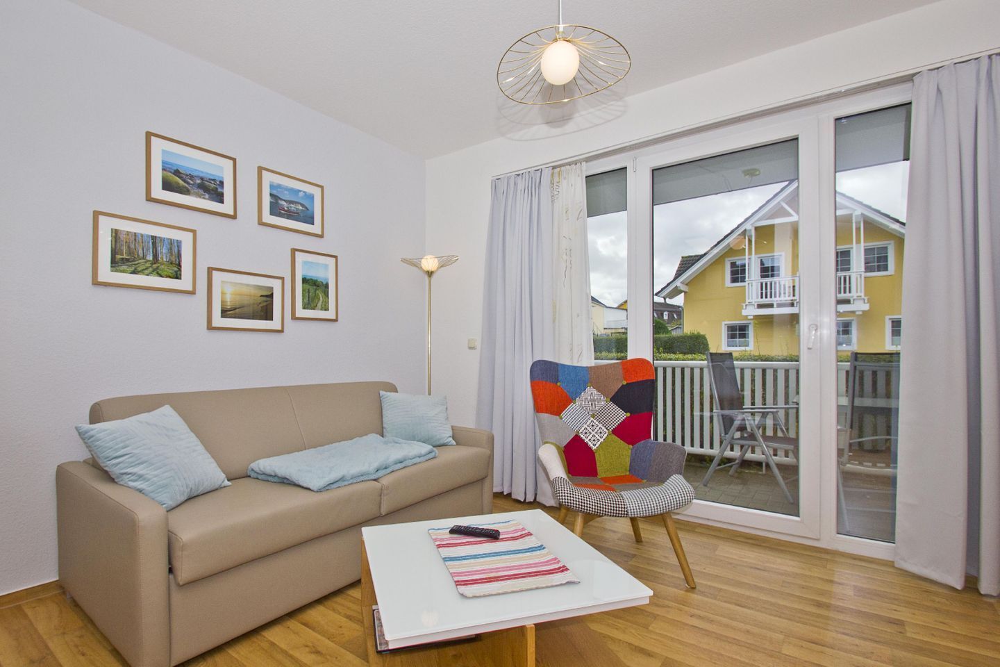 Un salon avec un canapé beige, un fauteuil coloré, une table basse avec une télécommande, et une grande fenêtre donnant sur un immeuble jaune.