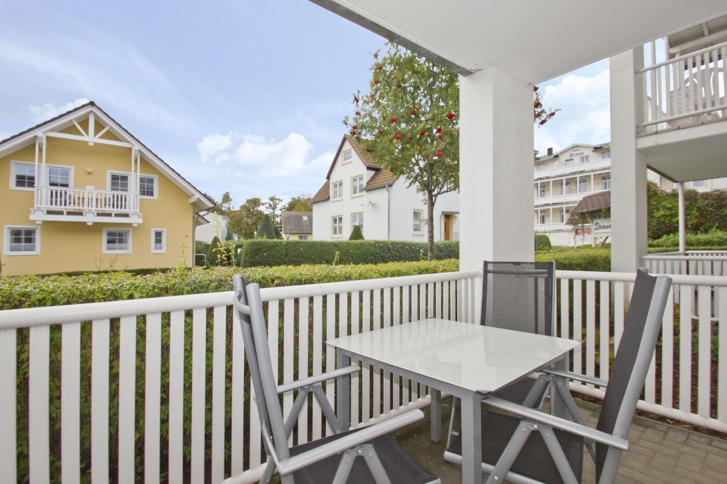 A covered outdoor area with a table and chairs, surrounded by white railings, in a residential neighborhood with houses and a tree with red berries.