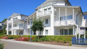 Un hôtel à plusieurs étages doté de balcons, un jardin parsemé de fleurs roses et un ciel d’un bleu limpide.