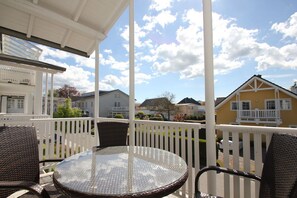A covered outdoor seating area with a round glass table and wicker chairs, overlooking a residential neighborhood with houses and a clear sky. - Balcony, free use of the AHOI adventure pool and sauna - Villa Karola FeWo 12 - (Göhren)