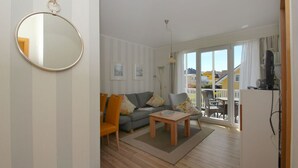 A living room with a sofa, chairs, a coffee table, and a TV. There is a balcony with a view of houses and a white ceiling. - Balcony, free use of the AHOI adventure pool and sauna - Villa Karola FeWo 12 - (Göhren)