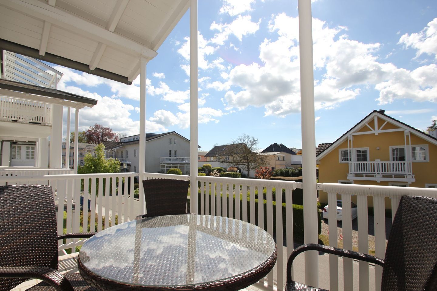 A covered outdoor seating area with a round glass table and wicker chairs, overlooking a residential neighborhood with houses and a clear sky.