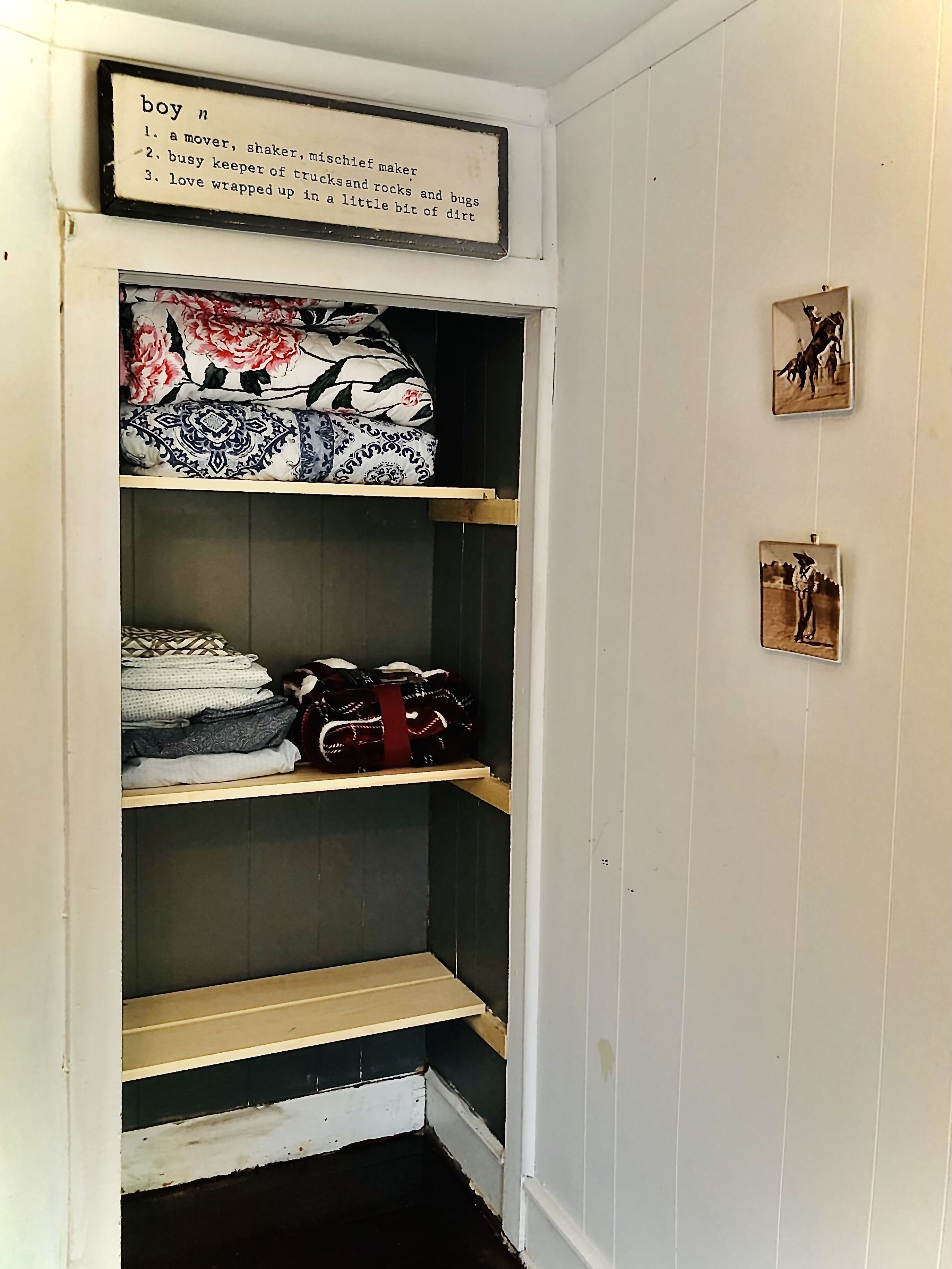 A closet with shelves containing folded clothes and bags, a framed sign with text, and two small framed pictures on the wall.