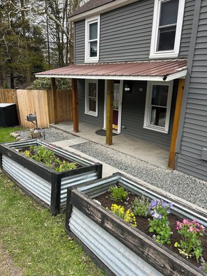 A house with a covered patio, a wooden deck, and raised garden beds with various plants. - Charming 120-year-old farmhouse by the creek (Hancock)