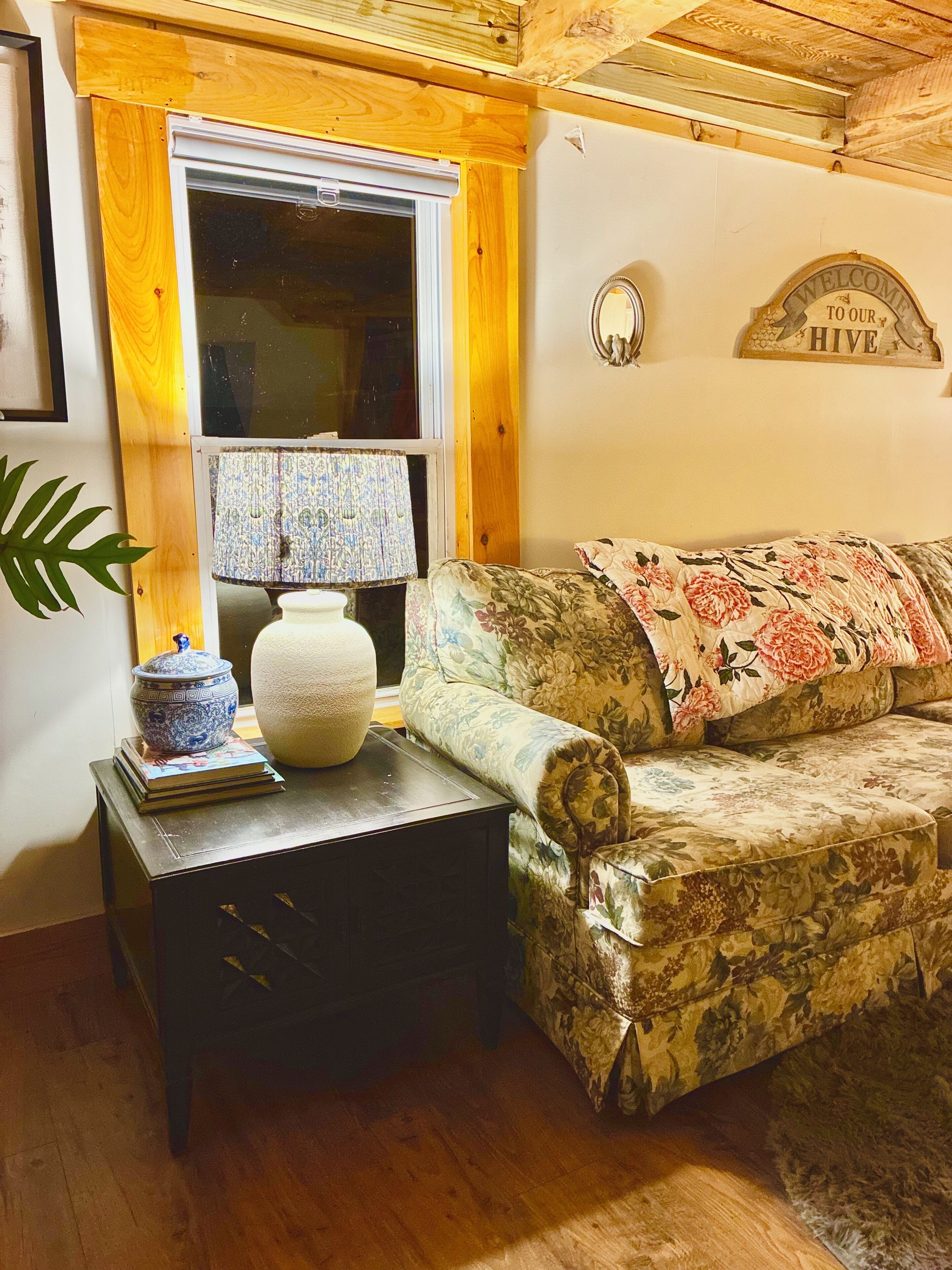 A cozy living room with a floral sofa, a small table with a lamp, a decorative bowl, and a welcome sign.