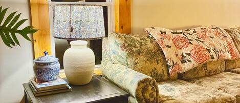 A cozy living room with a floral sofa, a small table with a lamp, a decorative bowl, and a welcome sign.